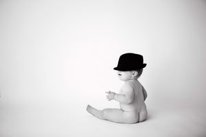 Black and white portrait of a baby wearing a black hat sitting against a light background.