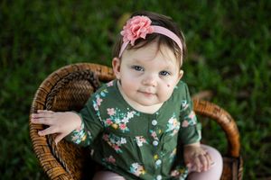 Baby in floral outfit sitting in wicker chair outdoors.