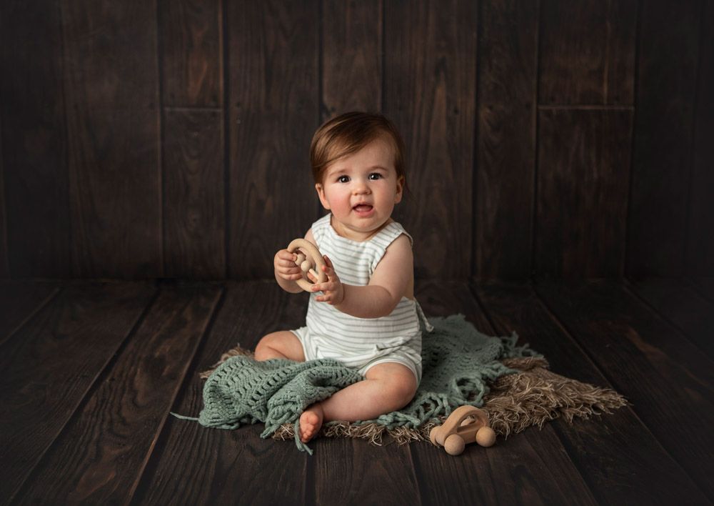 Baby in white outfit sits on green blanket against dark wooden backdrop with toys nearby.
