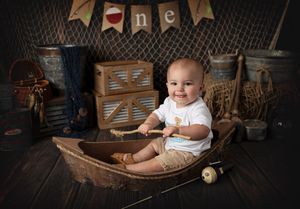 Baby sits in wooden boat prop with vintage decor and 'one' banner setting.