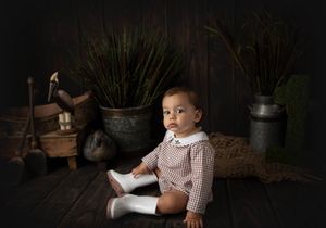 Baby in checkered outfit sits among vintage props and metal milk cans.