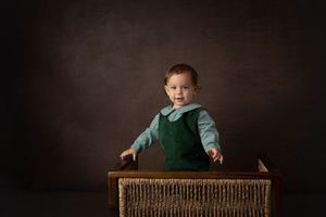 Baby in green overalls stands in wicker basket against dark backdrop.
