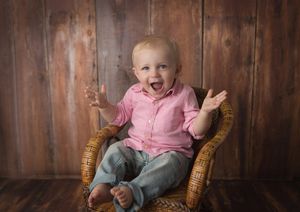 Sweet moment captured of baby in pink shirt sitting in wicker chair against wooden wall.