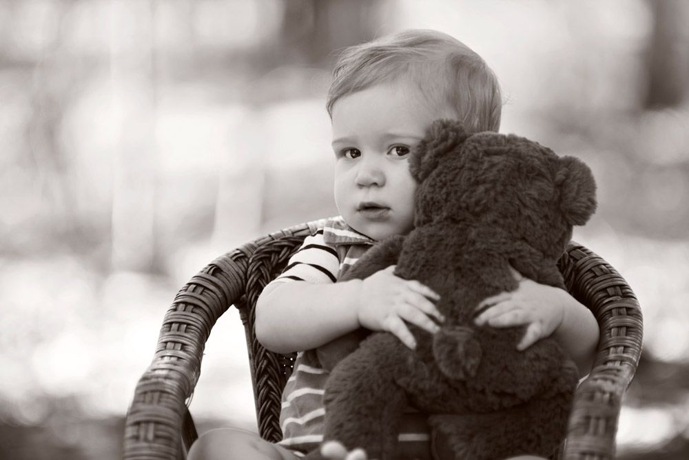 Black and white portrait of young child hugging teddy bear in wicker chair.