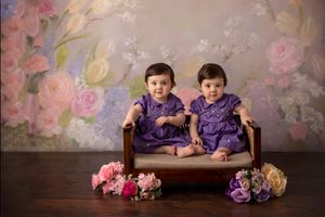 Two children in matching purple outfits sit on bench with floral backdrop.