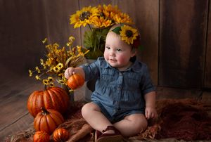 Baby in denim outfit sits with pumpkins and sunflowers in fall themed portrait.