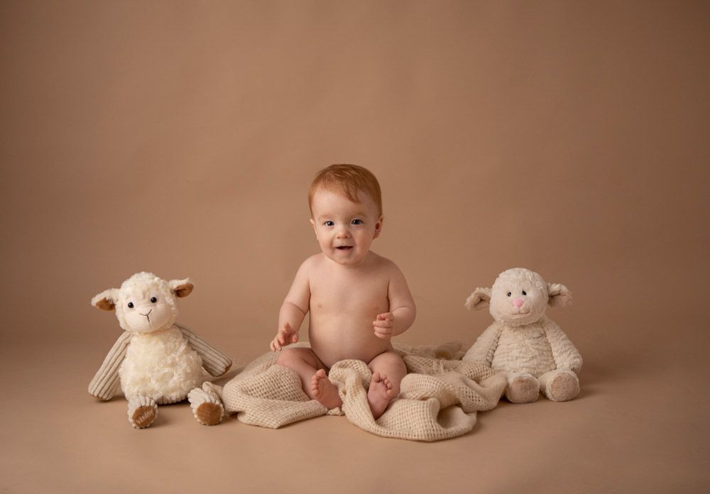 Baby in diaper sits between two plush lamb stuffed animals on beige backdrop.