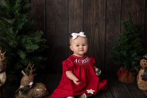 Holiday portrait with baby in red dress surrounded by Christmas trees and decor.