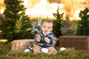 Baby in navy outfit sits on grass surrounded by evergreen trees and golden sunset.