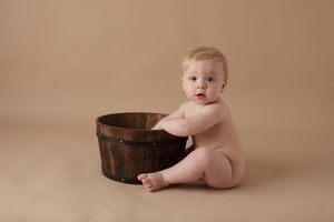 Baby sitting next to rustic wooden barrel on beige backdrop.