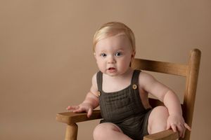 Baby wearing brown overalls sitting in wooden chair against neutral background.