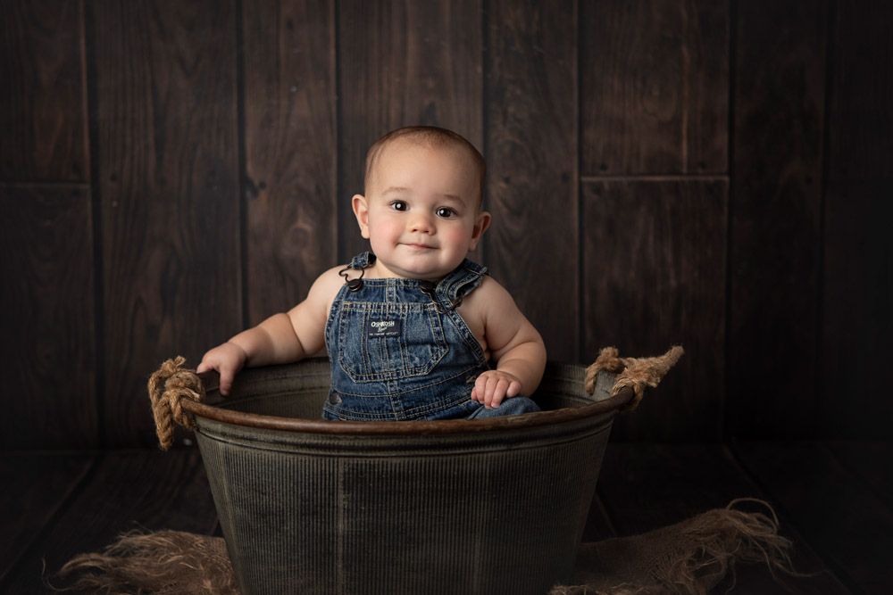 Baby in denim overalls sitting in metal washtub against dark wooden wall.