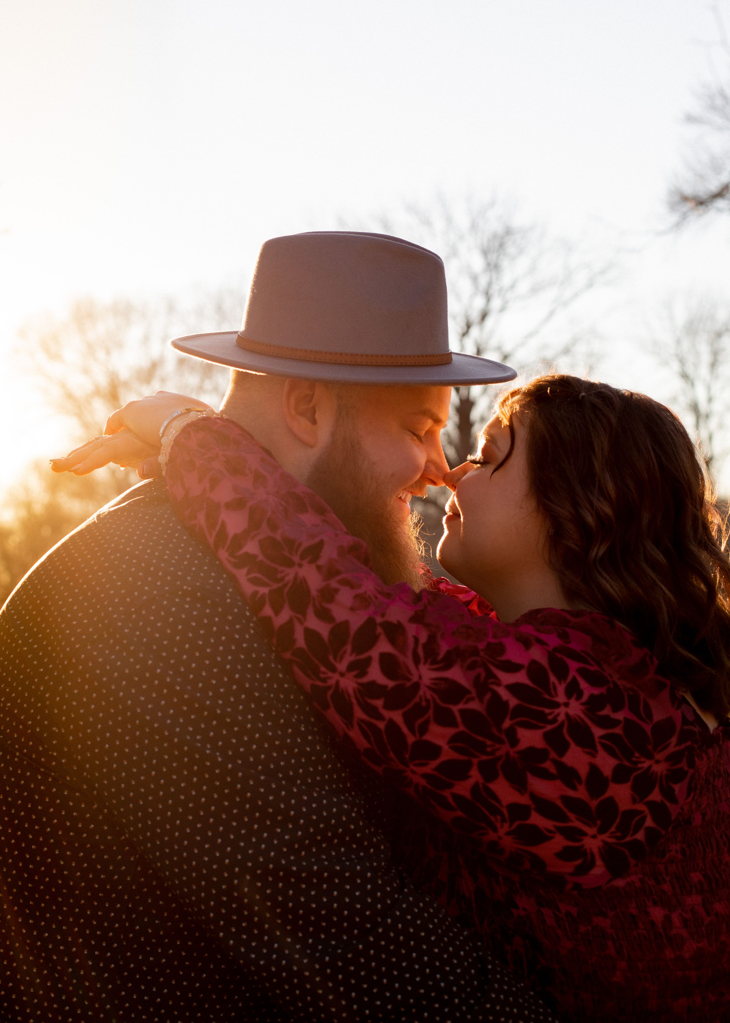 Silhouetted couple with hat share intimate moment during sunset in wooded setting.