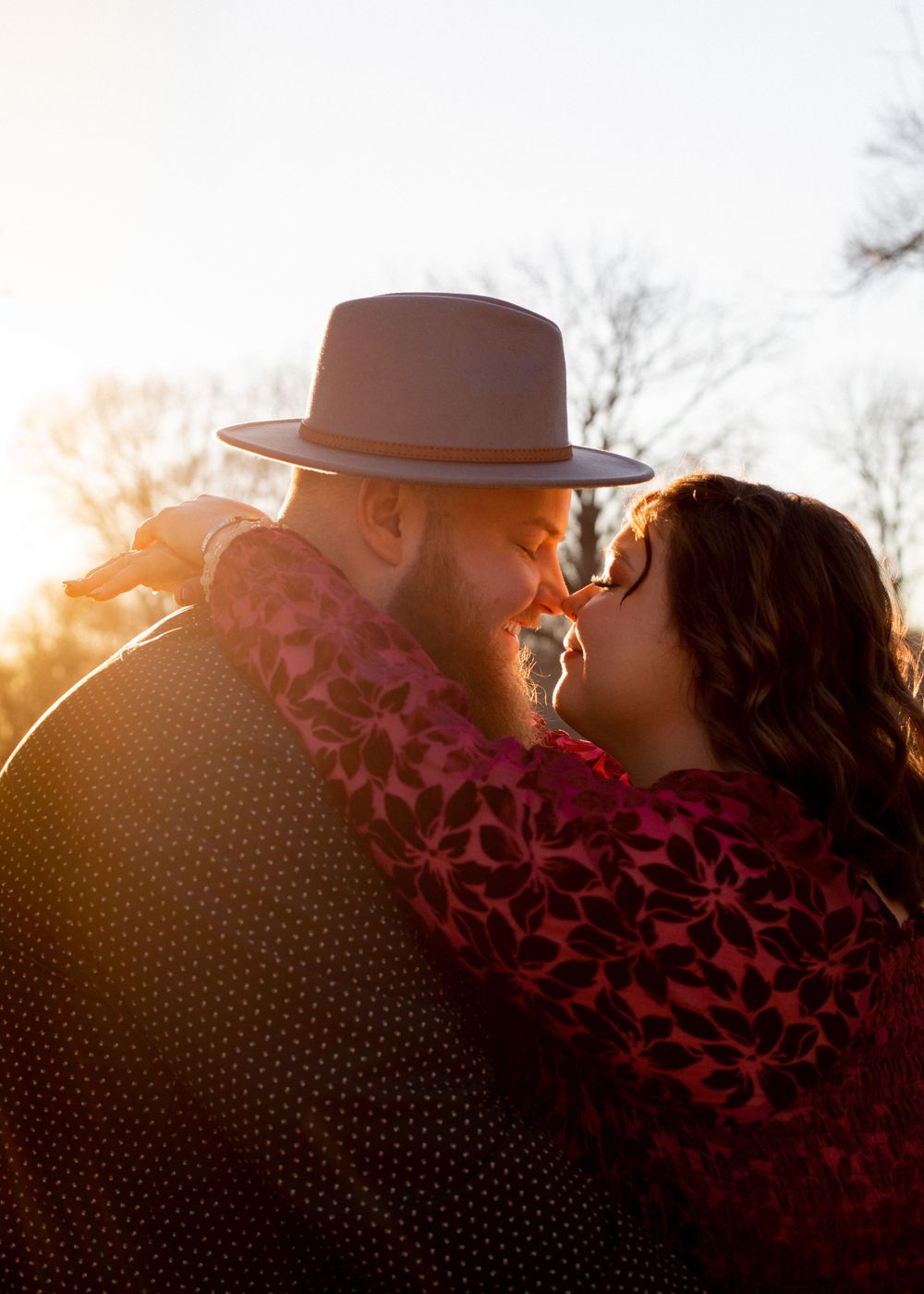 Silhouetted couple with hat share intimate moment during sunset in wooded setting.