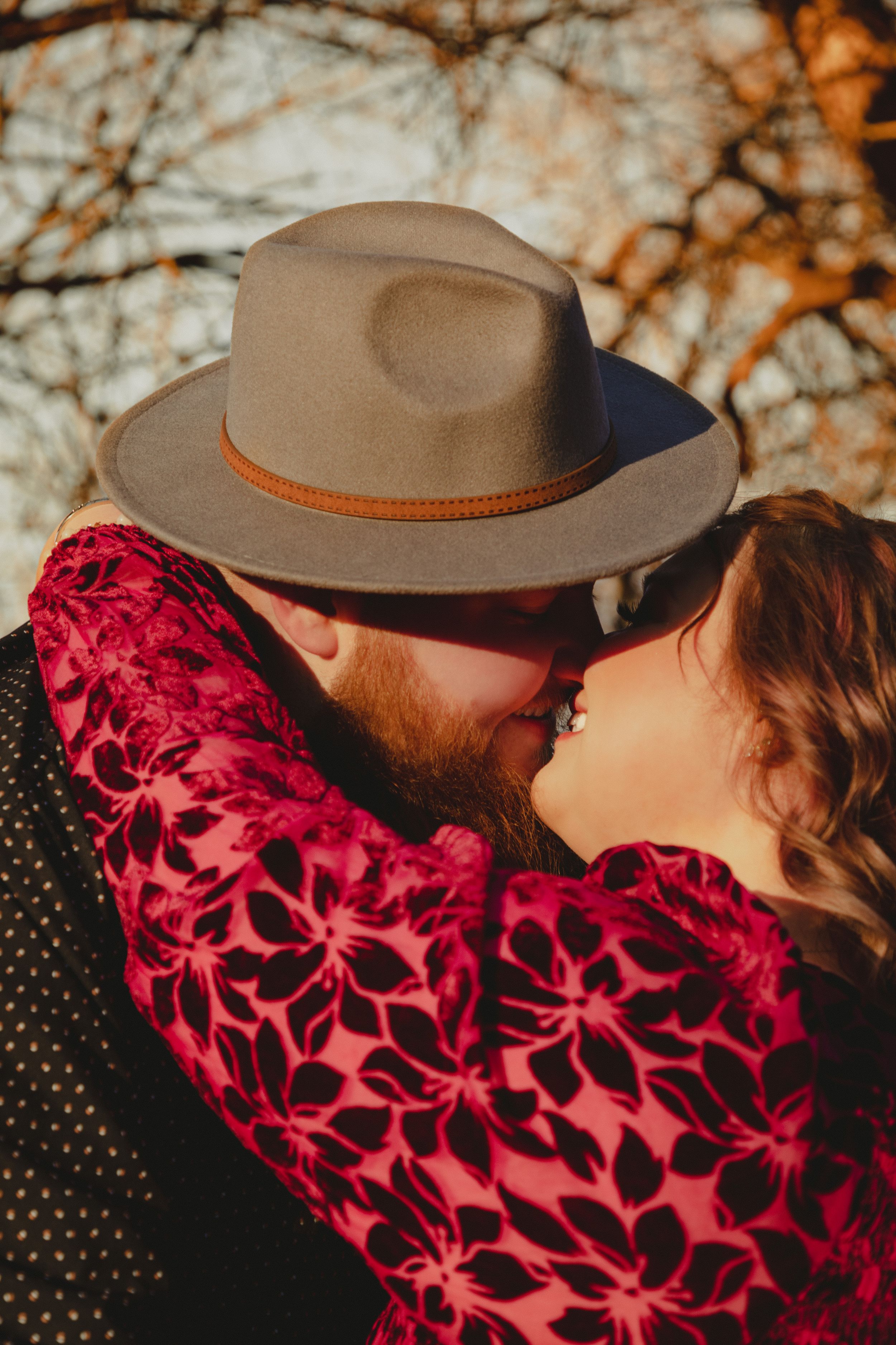 Romantic couple embracing in autumn light with person wearing brown hat and floral burgundy dress.