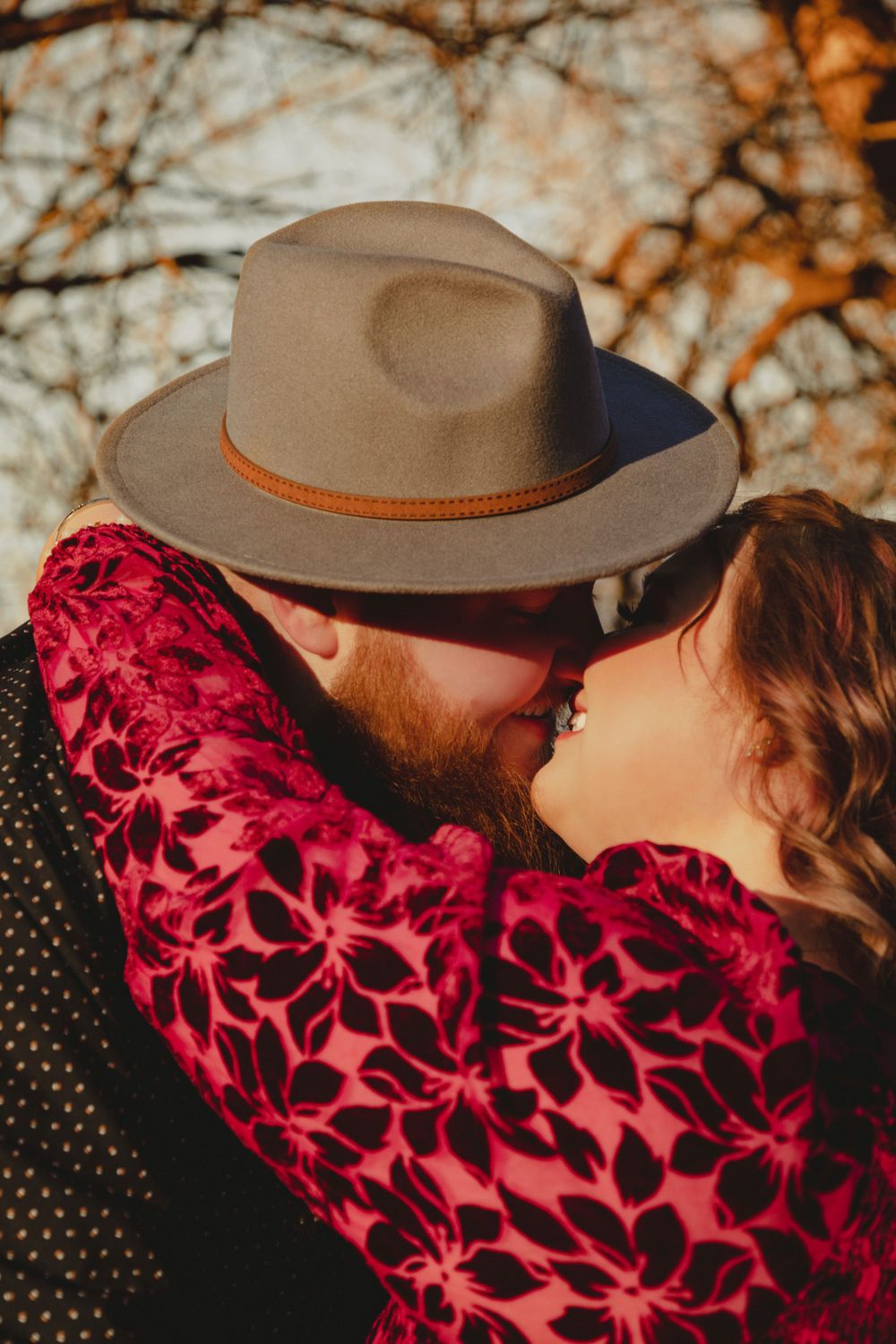Romantic couple embracing in autumn light with person wearing brown hat and floral burgundy dress.
