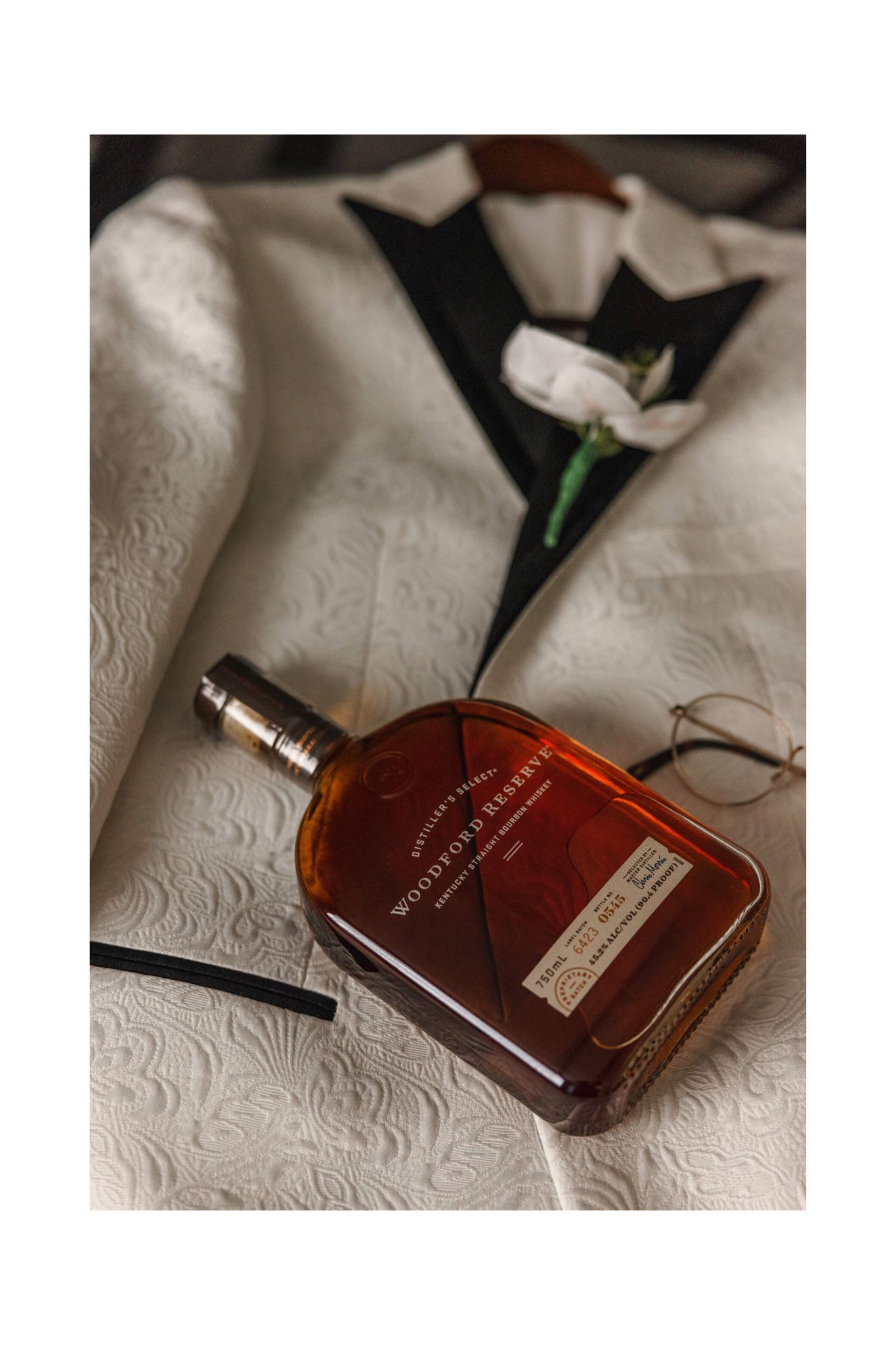 A bottle of whiskey rests beside formal wedding attire on a textured white surface.