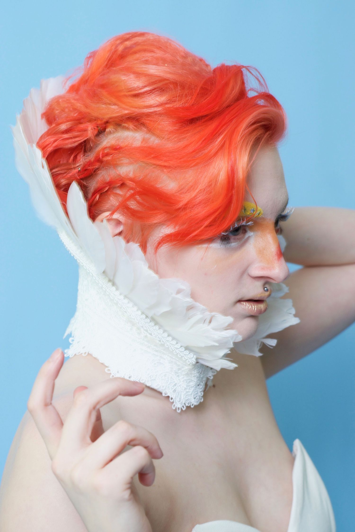 Avant garde beauty portrait featuring vibrant orange hair and white feather collar against blue background.