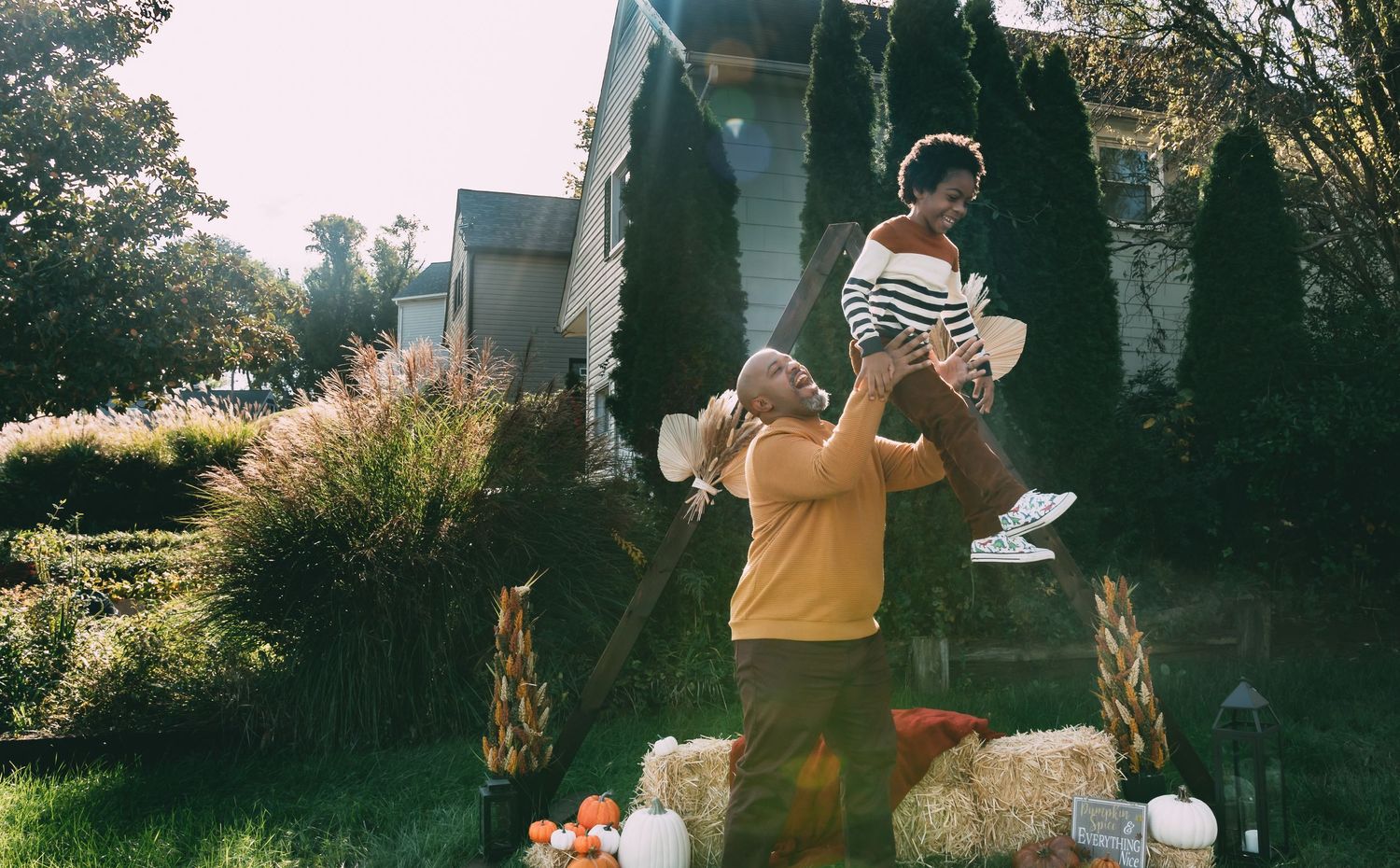 Father lifts smiling child into the air in a sunny backyard decorated with pumpkins and fall decor.