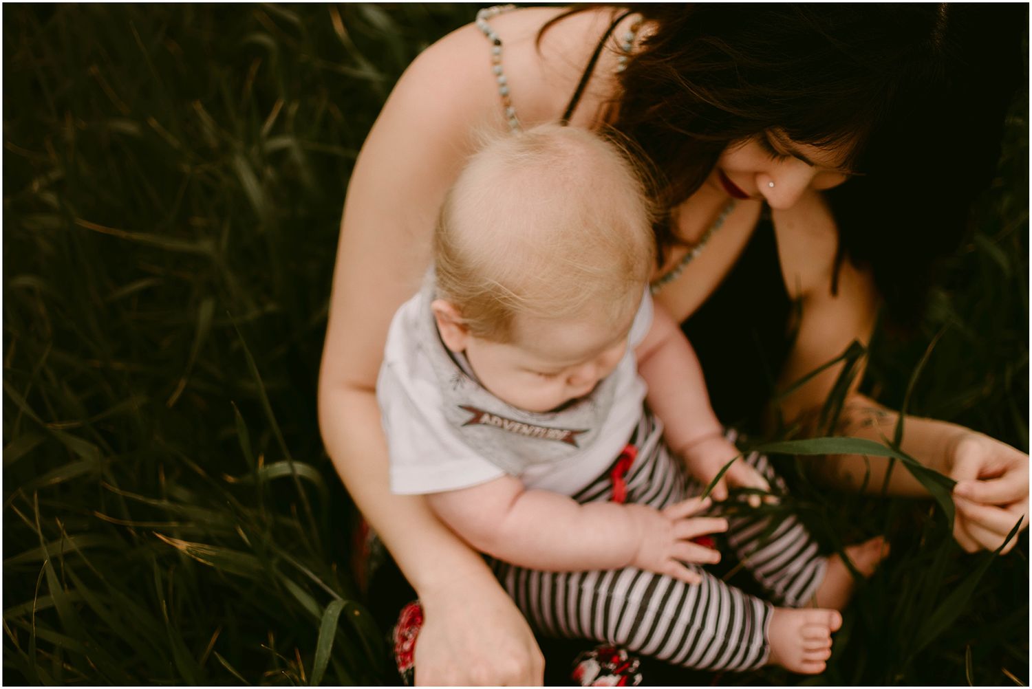 Baby in striped pants explores their surroundings while sitting with caregiver in tall grass.