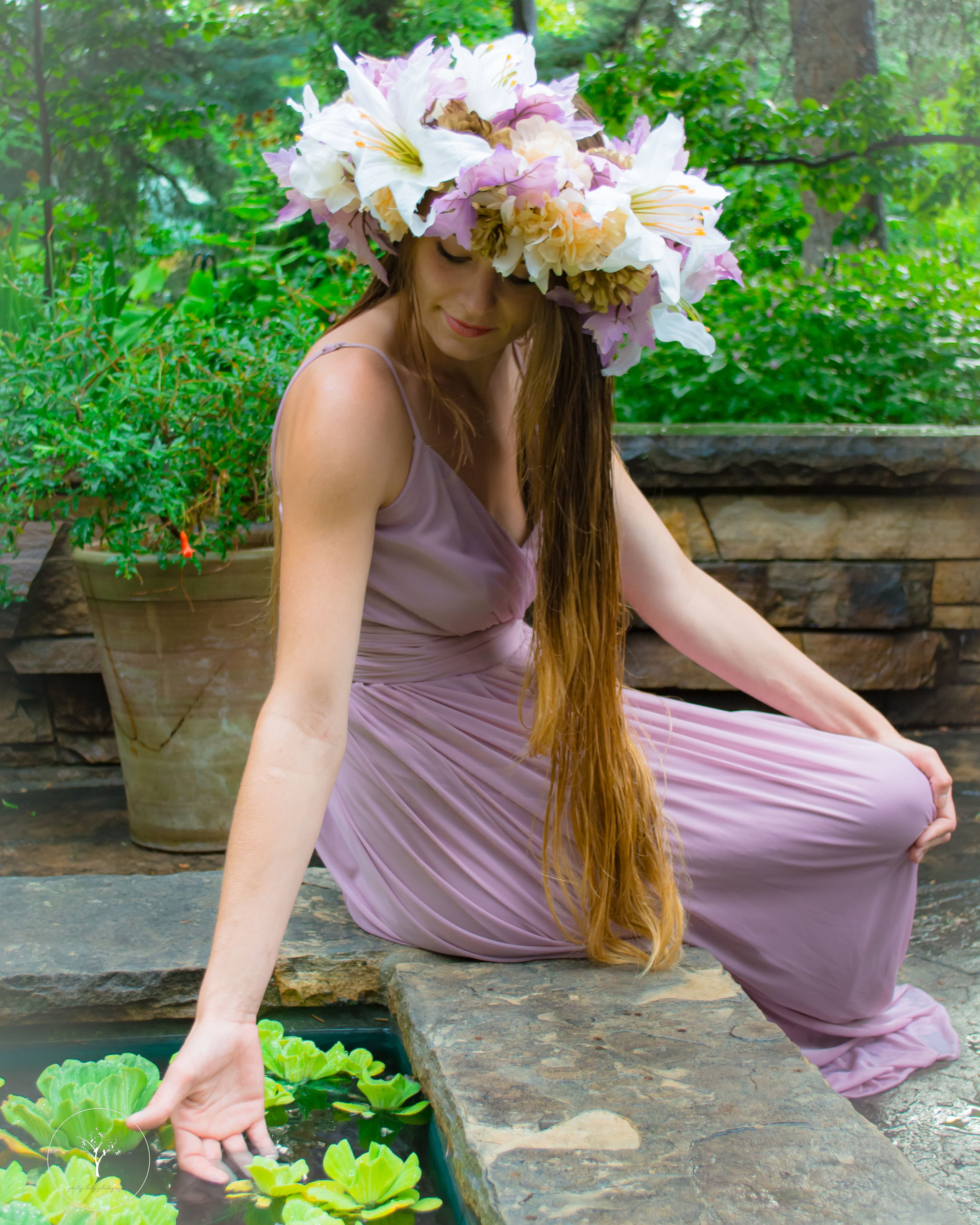 Person in flowing mauve dress sitting on stone wall surrounded by greenery.
