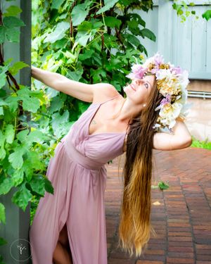Woman in flowing pink dress poses dramatically with flowers in her hair against ivy-covered wall.
