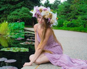 Ethereal portrait of woman in lavender dress with floral crown sitting by lily pond.