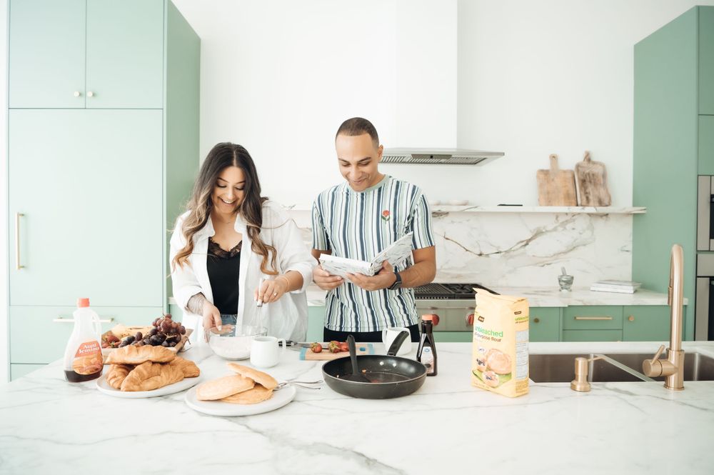 Couple cooking together in a modern mint green kitchen, preparing a meal with pastries and ingredients on marble counter.