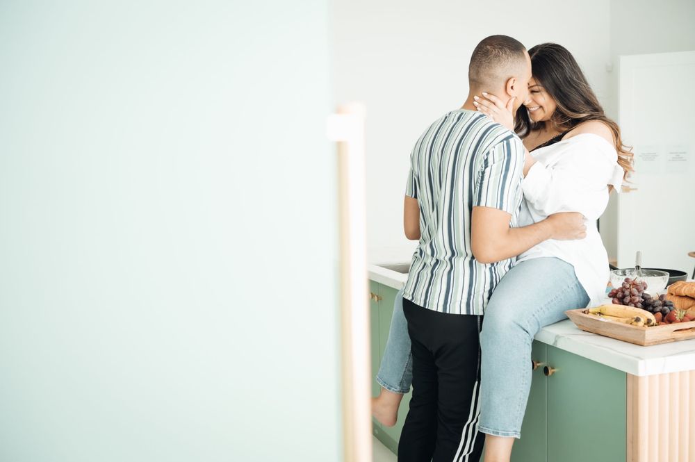 Romantic couple embracing in a stylish mint green kitchen, woman sitting on counter while partner holds her close.