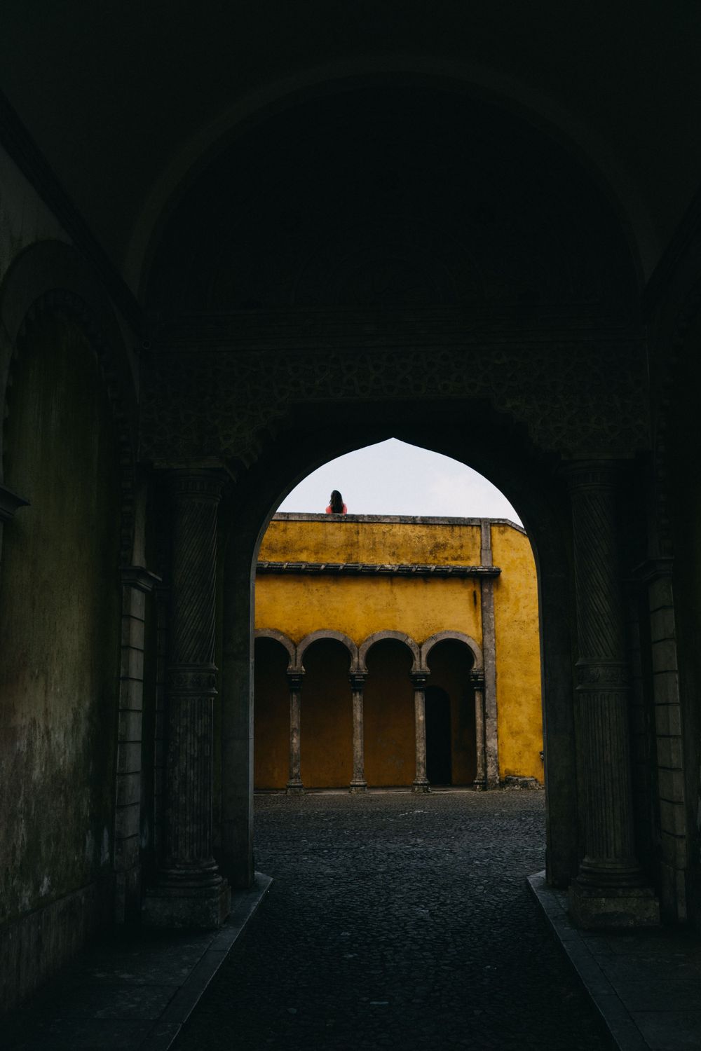 Dark archways in a historic building lead to a sunlit yellow courtyard.