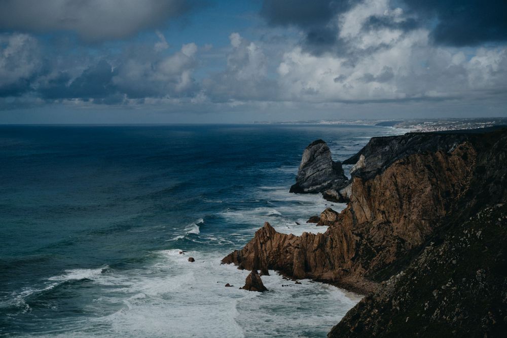 Dramatic rocky coastline with crashing waves meets dark cloudy skies over the ocean.