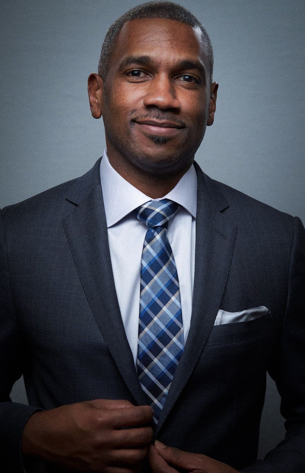 Professional Black man in dark suit with blue plaid tie and pocket square smiling confidently.