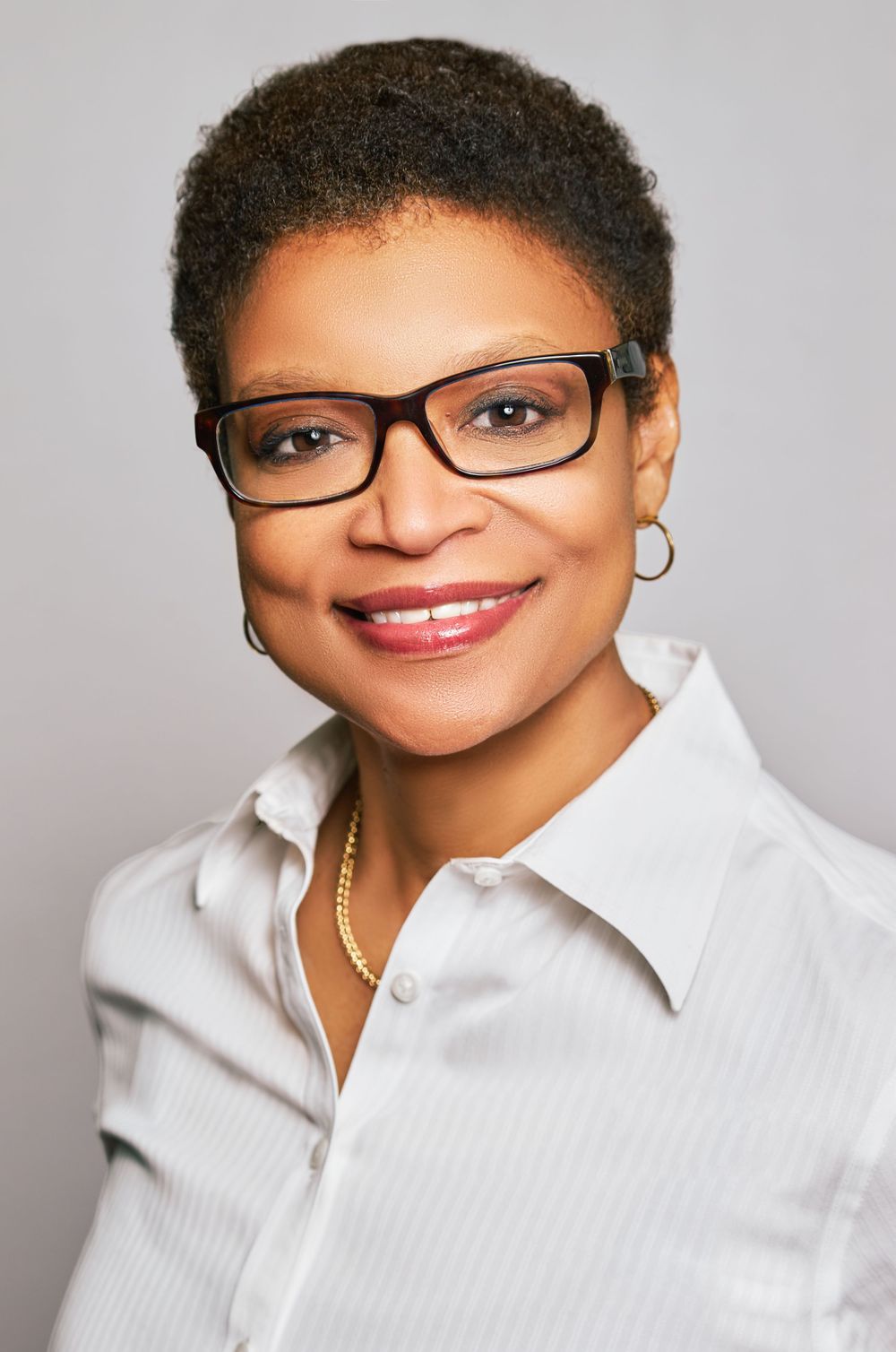 Professional headshot of a smiling Black woman wearing glasses, gold jewelry, and a white button-up shirt against a gray background.