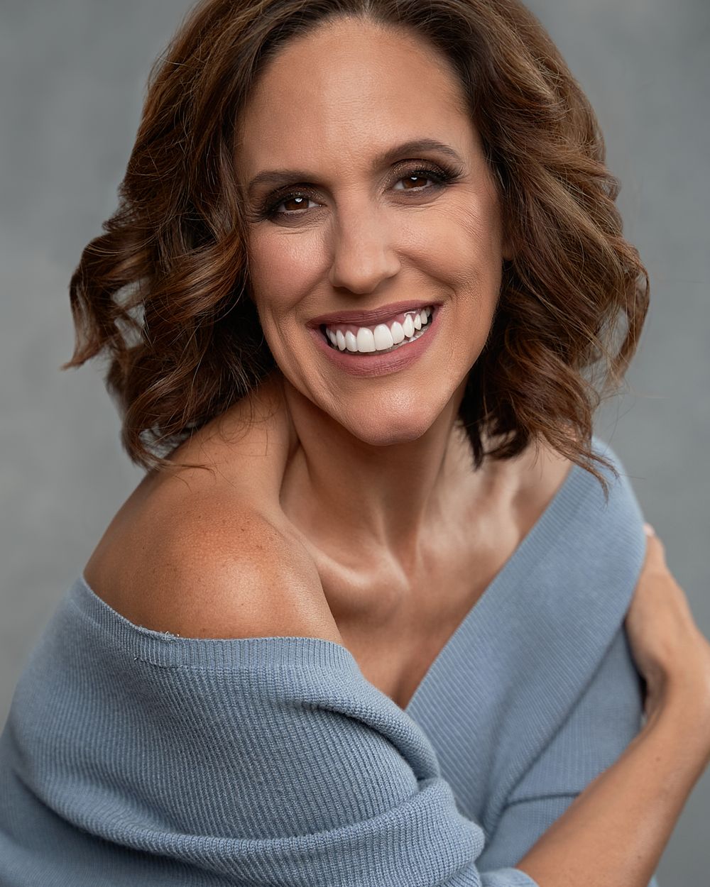 Smiling middle-aged woman in off-shoulder blue sweater posing for a professional headshot.