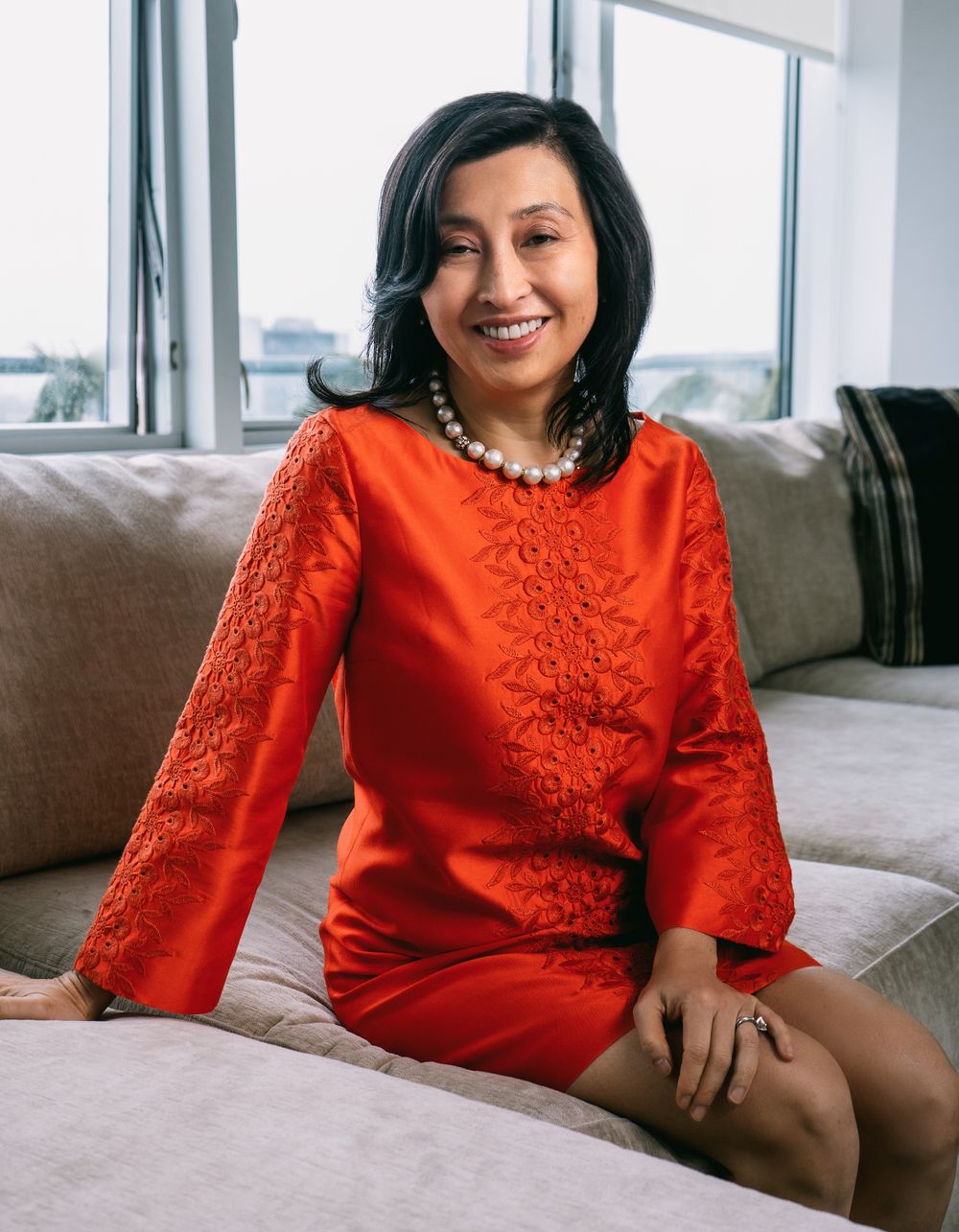 A smiling woman in a red lace long-sleeve dress sits on a sofa wearing a pearl necklace near bright windows.