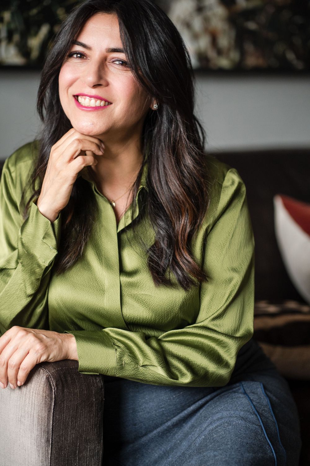Smiling woman in green satin blouse sits on sofa with hand on chin.