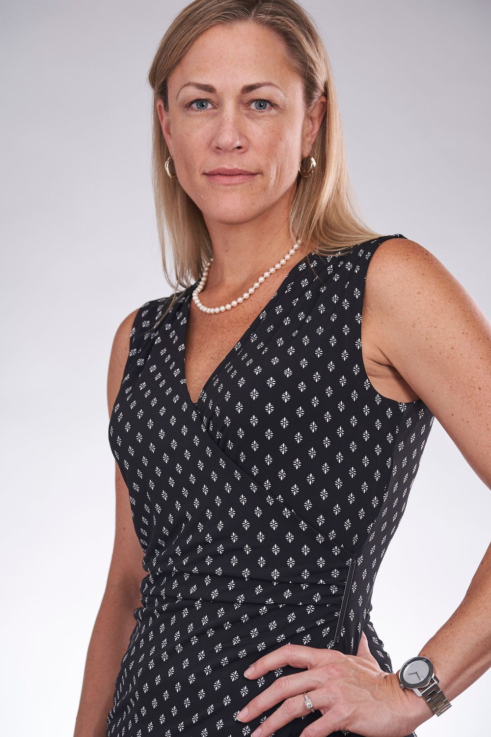 Professional blonde woman in black patterned dress with pearl necklace posing confidently.