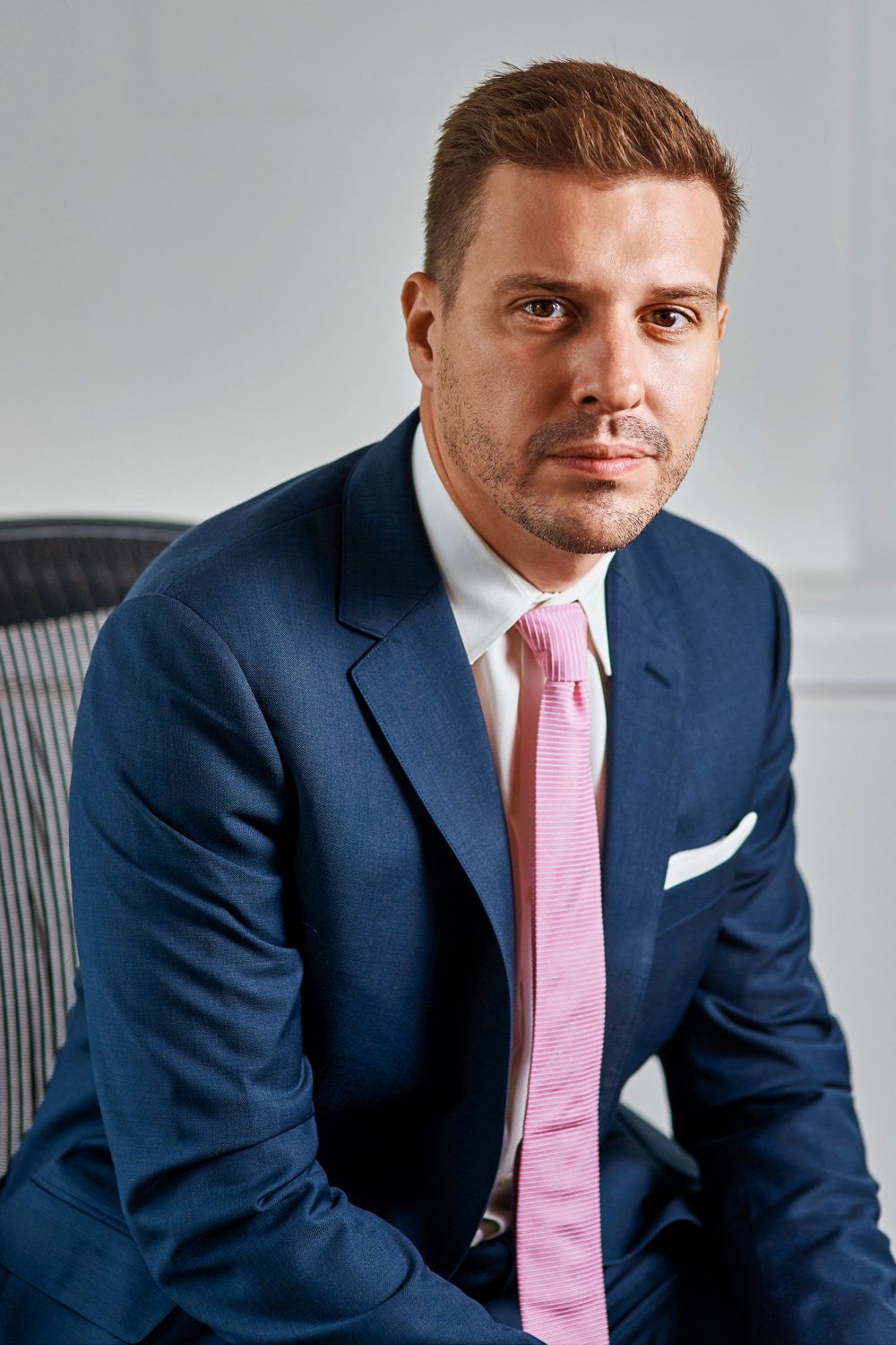 Professional man in navy suit and pink tie seated in office chair with serious expression.