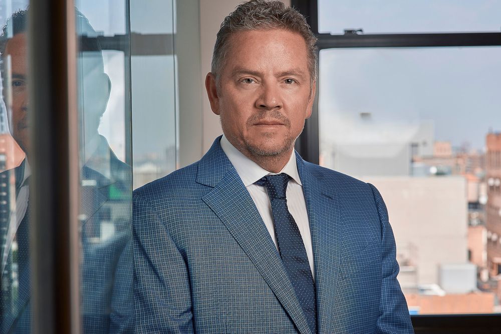 Professional man in blue suit standing near office window with city view behind him.