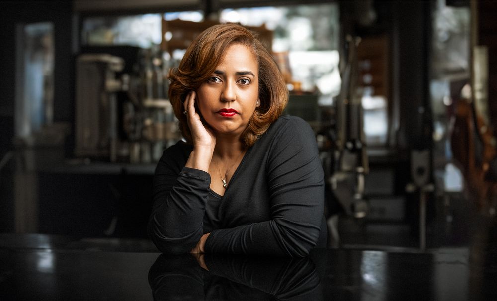 A woman in a black top rests her chin on her hand, posing thoughtfully at a reflective table in a dimly lit setting.