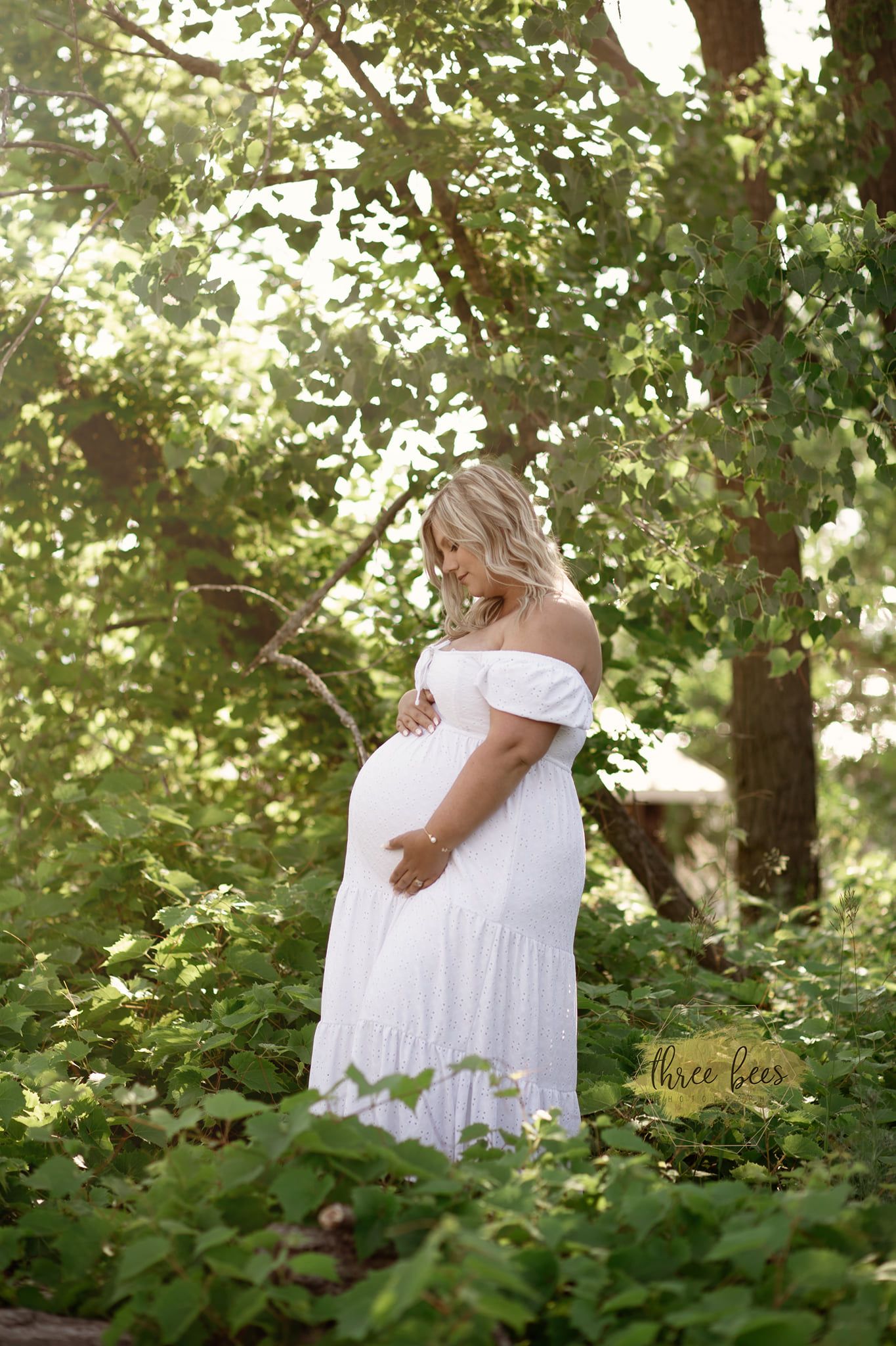 A maternity portrait of a woman in a white dress standing among green foliage.