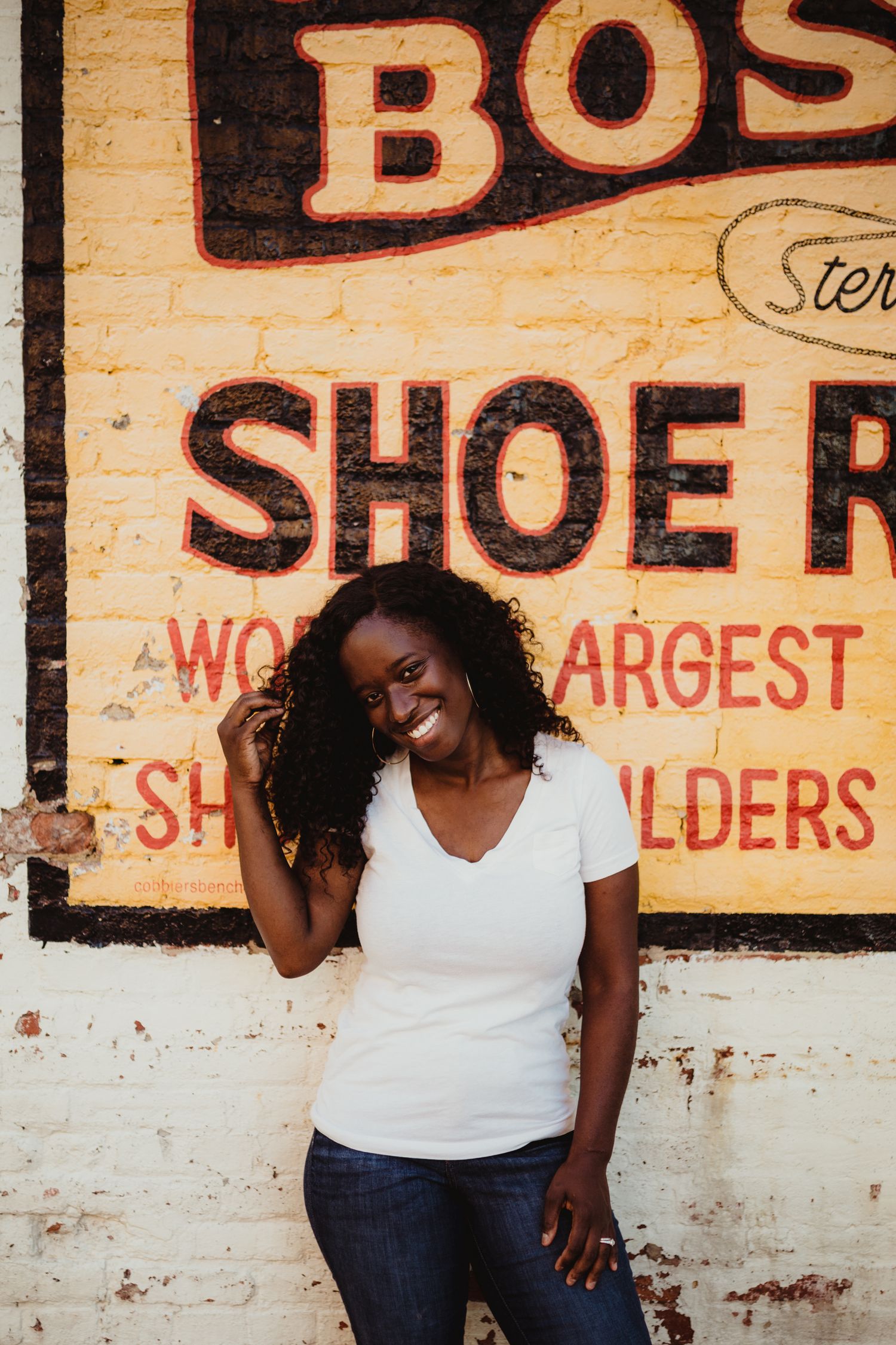 a woman smiles while leaning on a brick wall which has a chipped advertisement for shoe repair painted on it