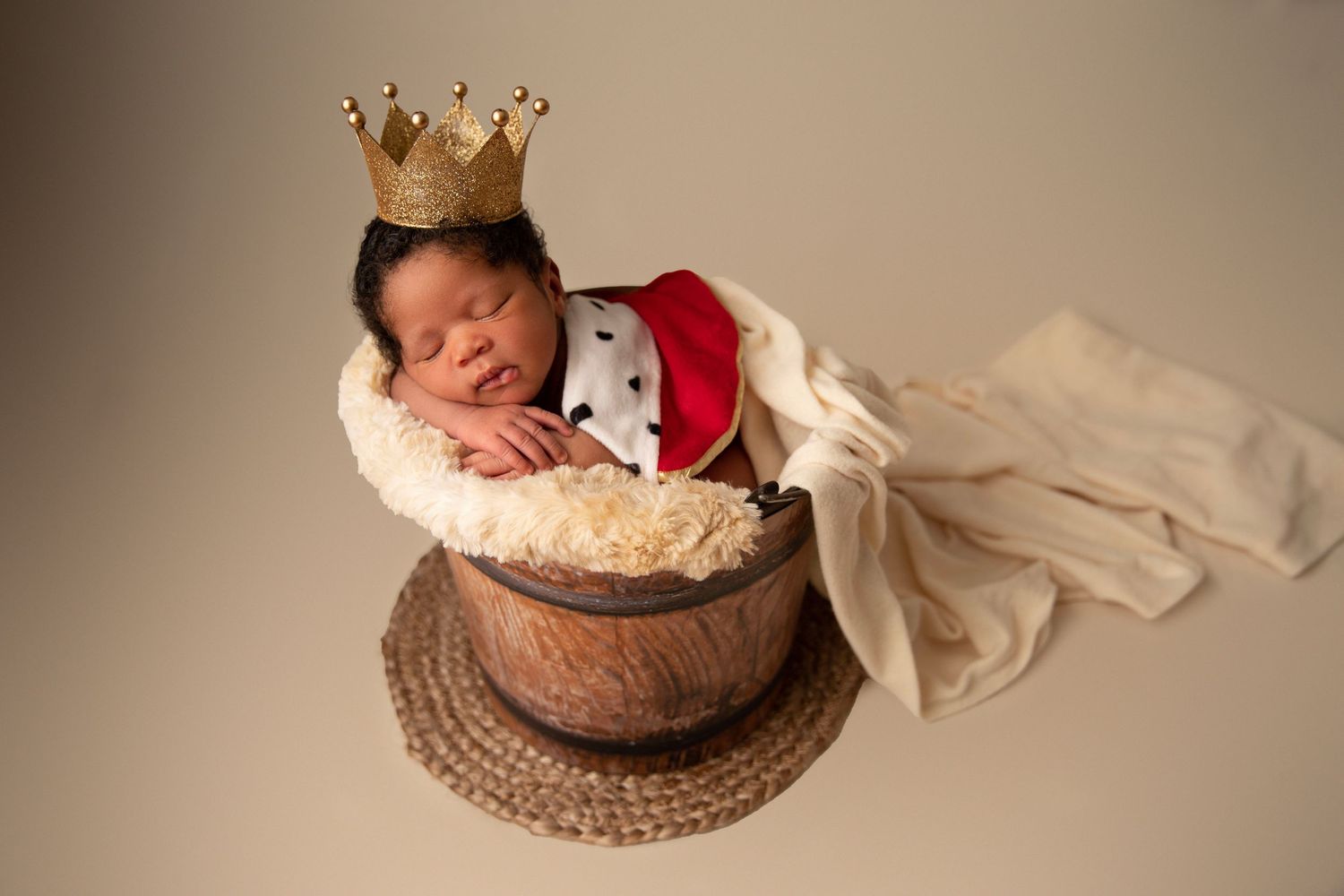 Newborn baby sleeping in wooden bucket wearing king costume with gold crown and red cape on neutral background.