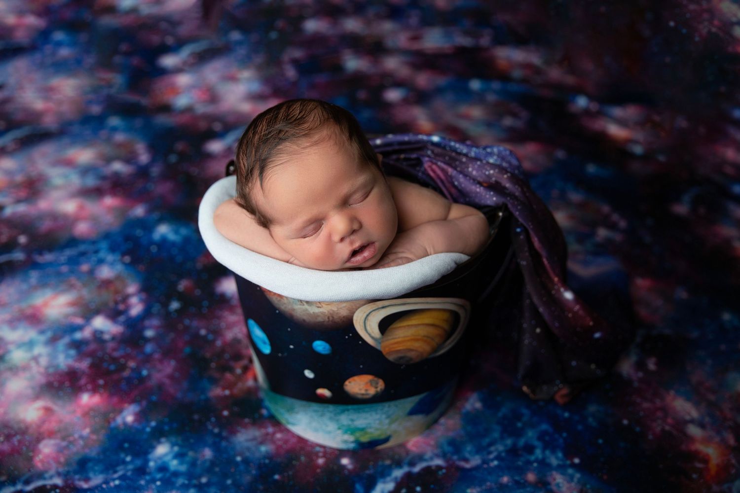 Newborn baby sleeping in space-themed painted bucket with galaxy backdrop and purple wrap.