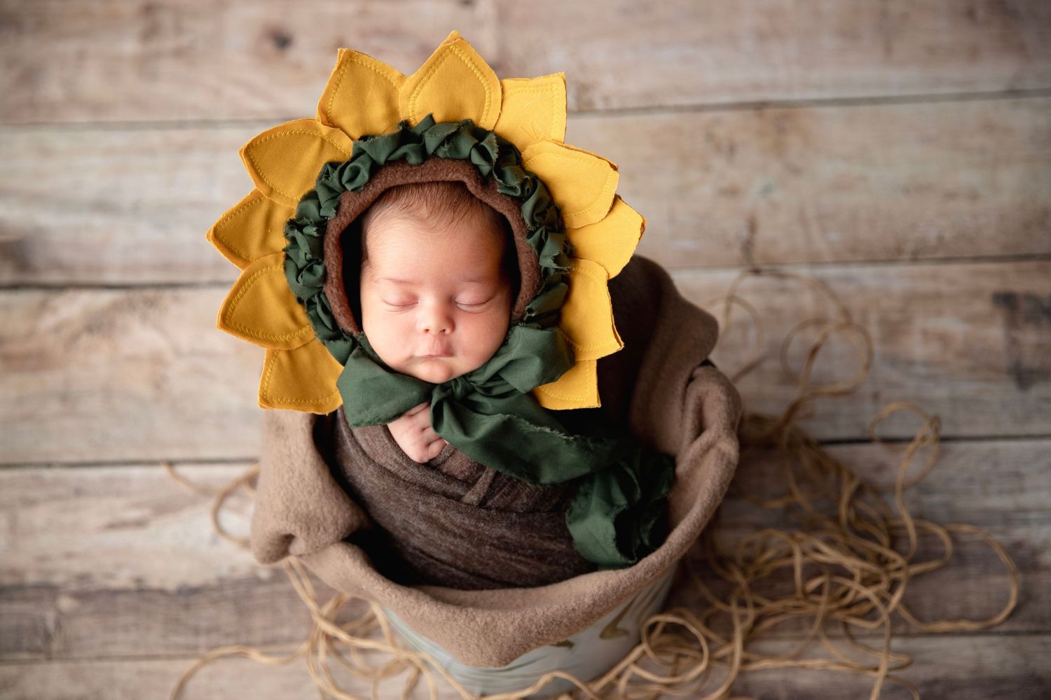Sleeping newborn baby wearing sunflower bonnet wrapped in brown swaddle in burlap pot on wood background.