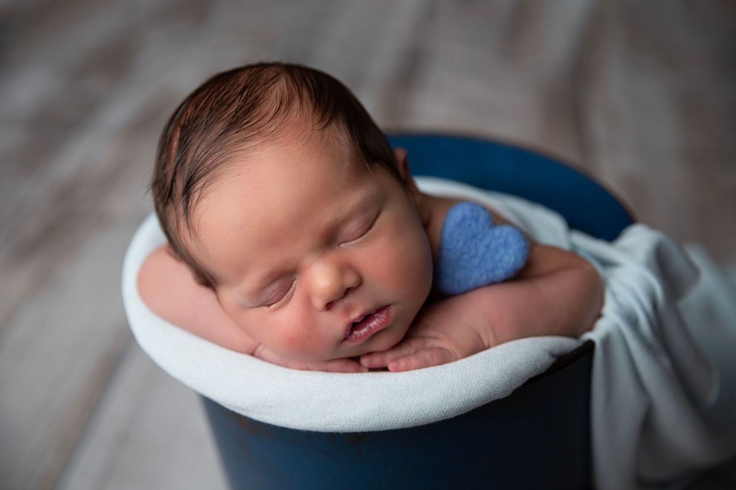 Sleeping newborn baby resting in blue bucket with soft blue heart accent on rustic wood background.