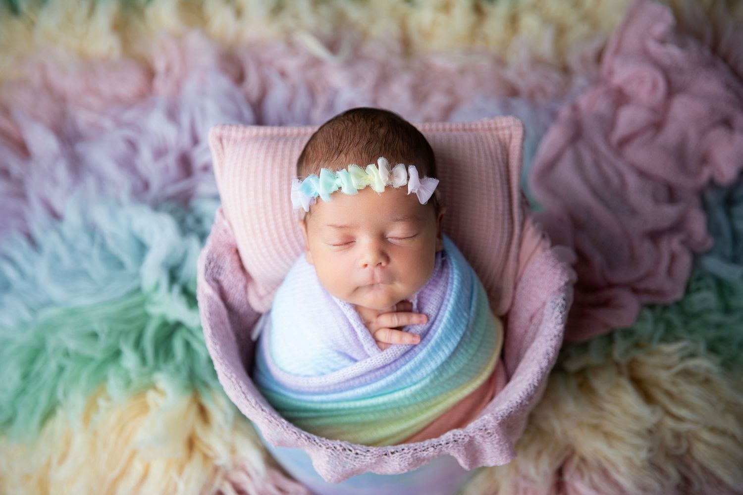 Sleeping newborn baby girl swaddled in rainbow wrap with floral headband on colorful fluffy rug.