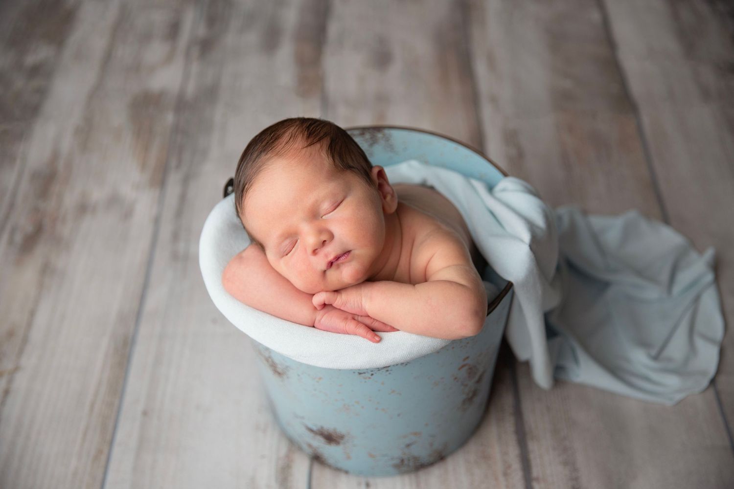 Peaceful newborn baby sleeping in rustic blue bucket with light blue wrap on wood floor.