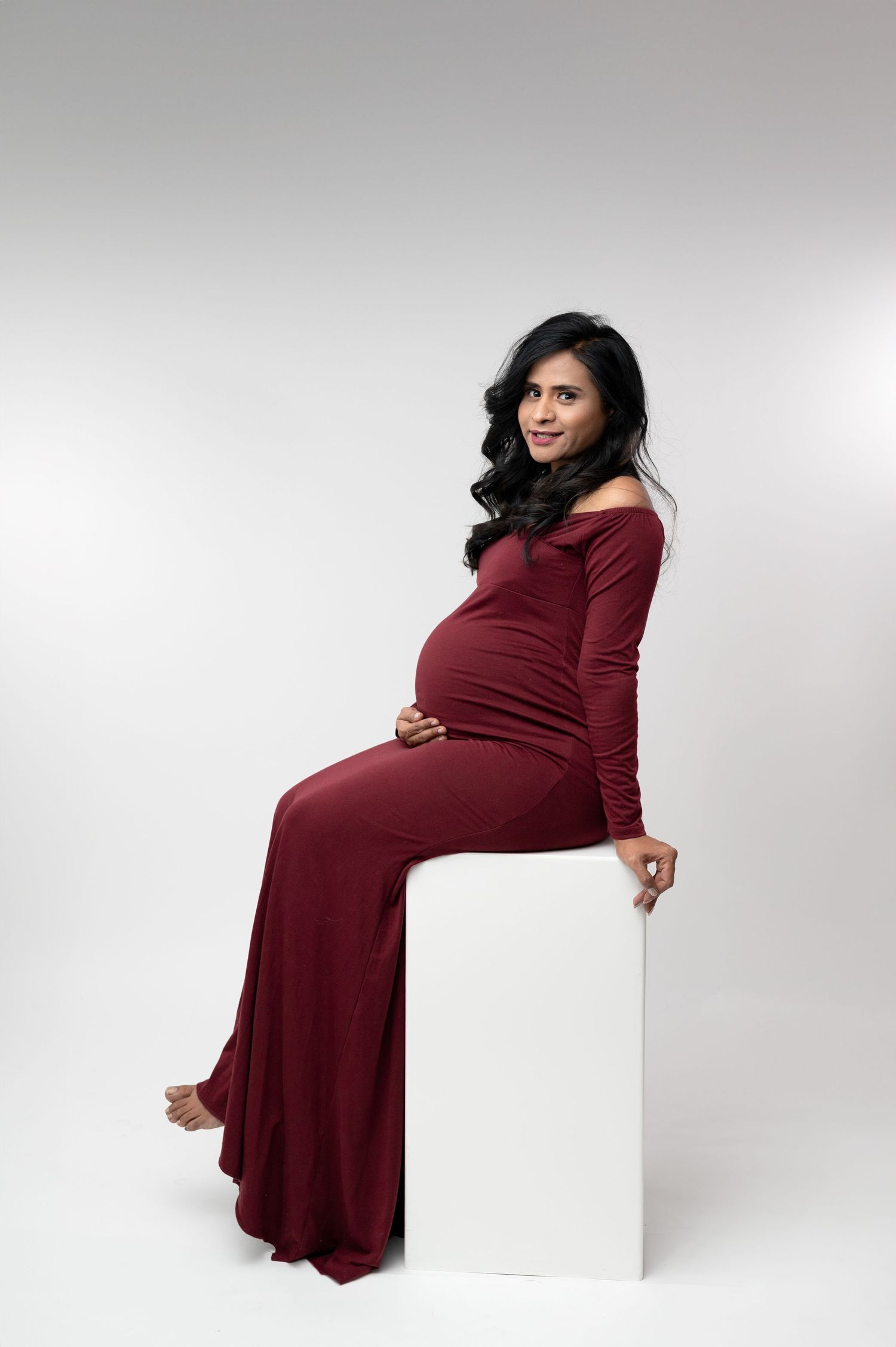 Expectant mother in burgundy dress poses elegantly against white backdrop.