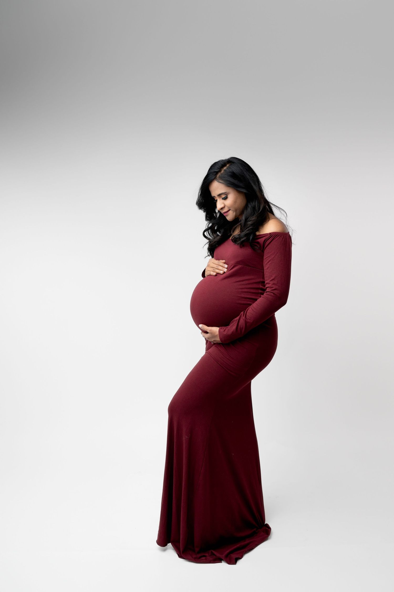Woman in burgundy dress cradles baby bump in artistic studio portraits.